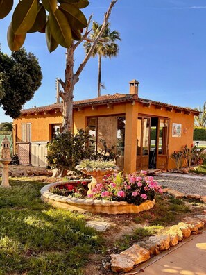 Ein eingeschossiges Haus mit Ziegeldach, einer Haustür und einem kleinen Garten mit Blumen und einem Brunnen.