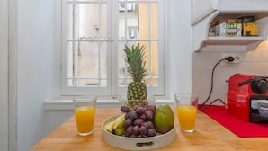Eine Küche mit einer Obstschale mit Trauben, einer Ananas und einem Mango, zwei Gläsern Orangensaft und einer roten Kaffeemaschine auf einem Holztisch.