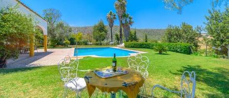A poolside area with a table and chairs, a bottle of wine, and a view of the pool and surrounding greenery.