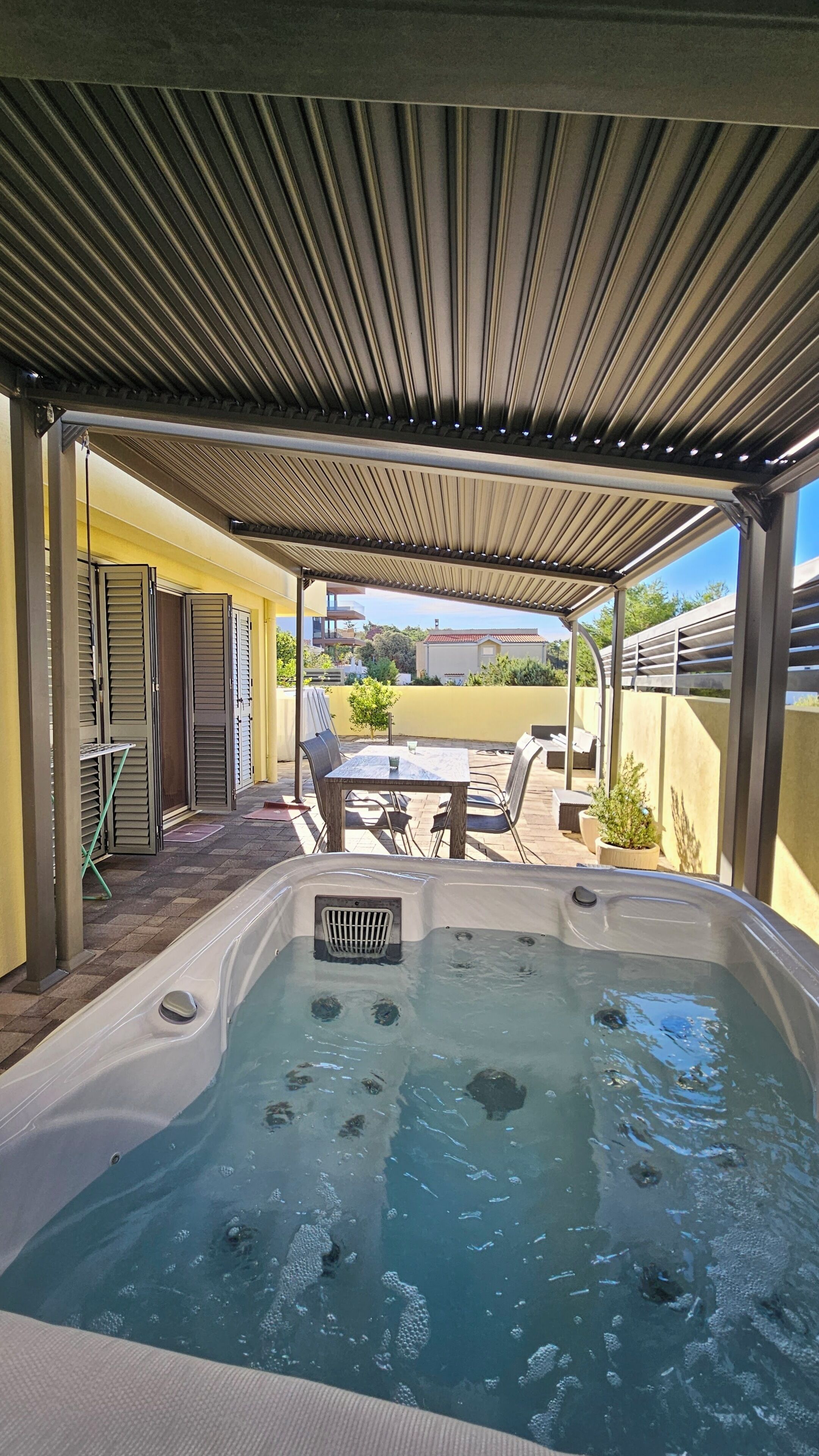 A hot tub under a pergola with a patio table and chairs, surrounded by yellow walls and a view of the neighborhood.