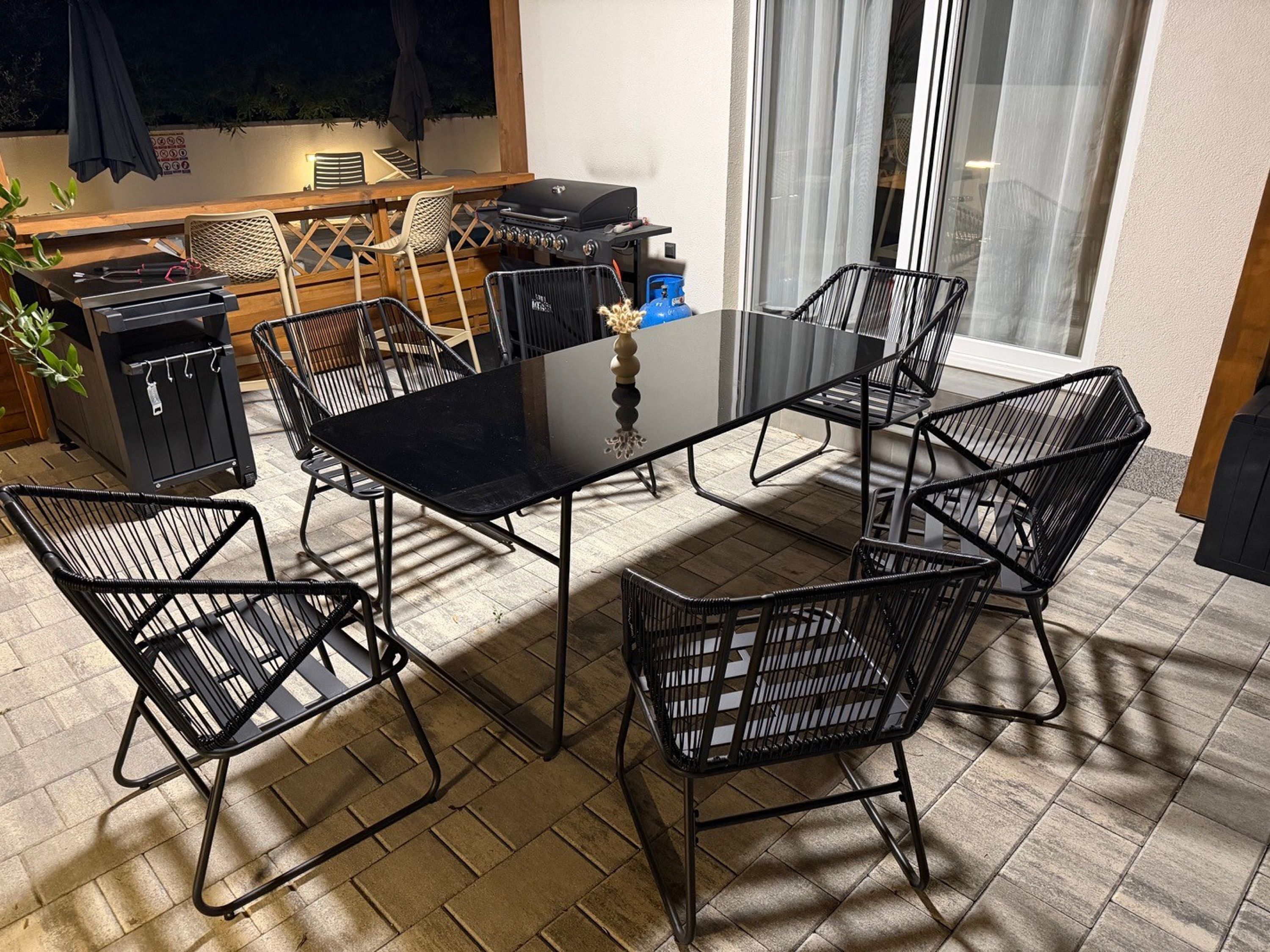 Une terrasse avec une table en verre entourée de chaises en métal, un barbecue et une poubelle.