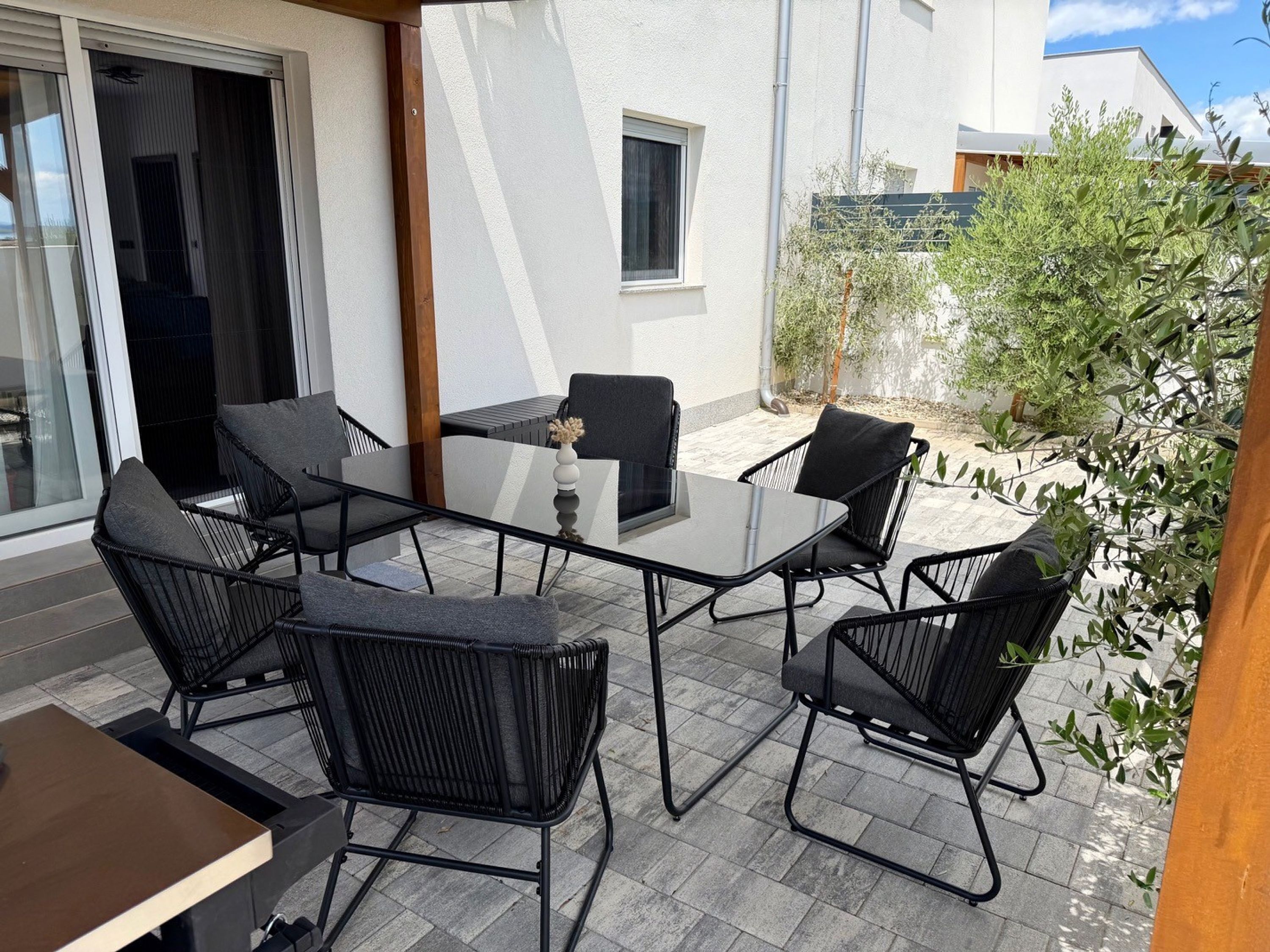 An outdoor patio with a glass table and four chairs, a small vase, and a view of a building and greenery.