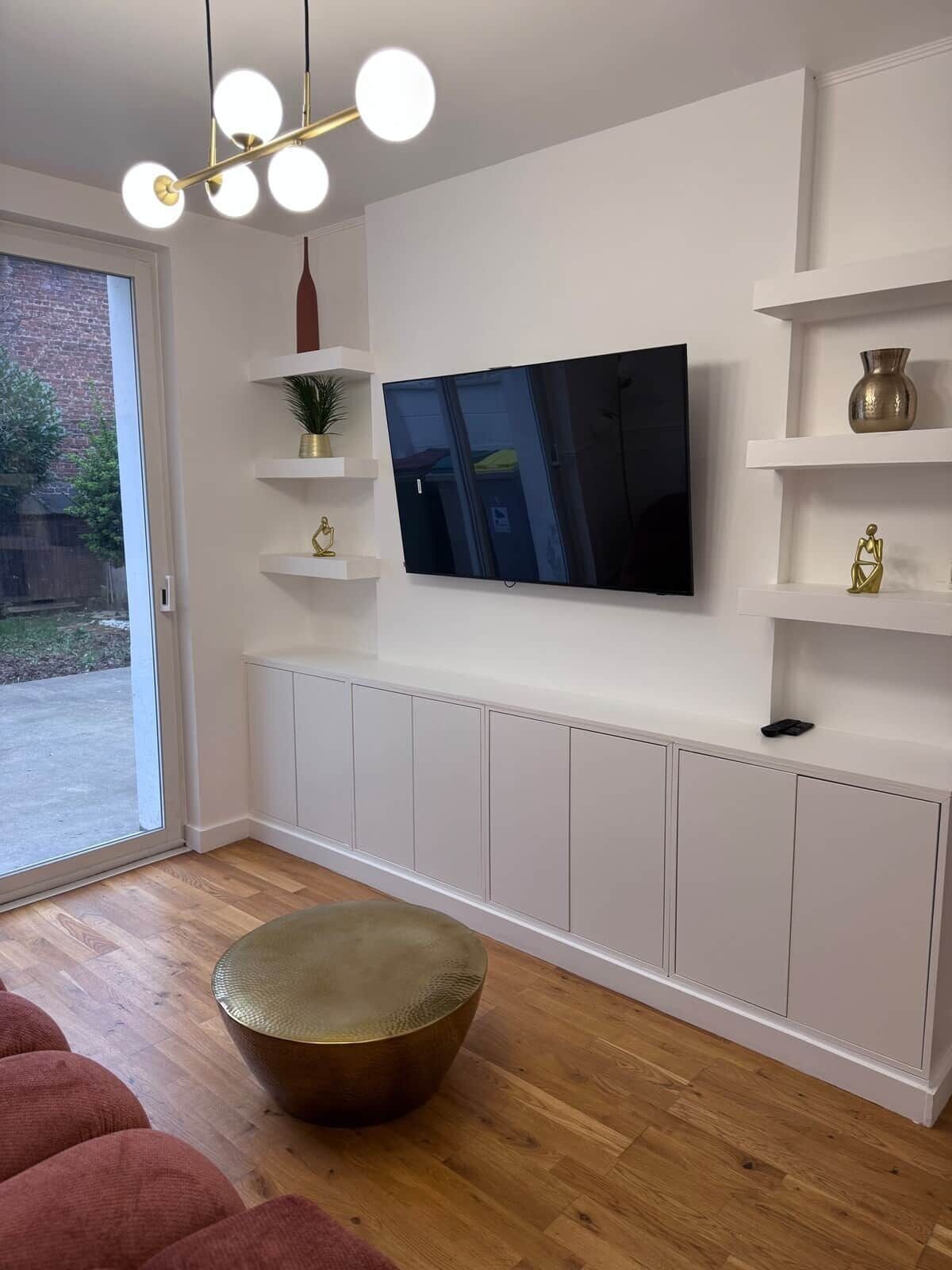 Ein modernes Wohnzimmer mit Flachbildfernseher an einer weißen Wand, Holzboden und einem runden Hocker.