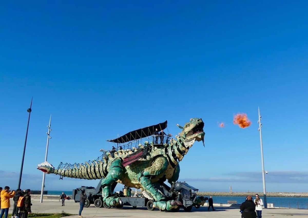Un grand dinosaure mécanique, avec une personne assise sur son dos, est installé sur une surface pavée, avec un ciel dégagé en arrière-plan.
