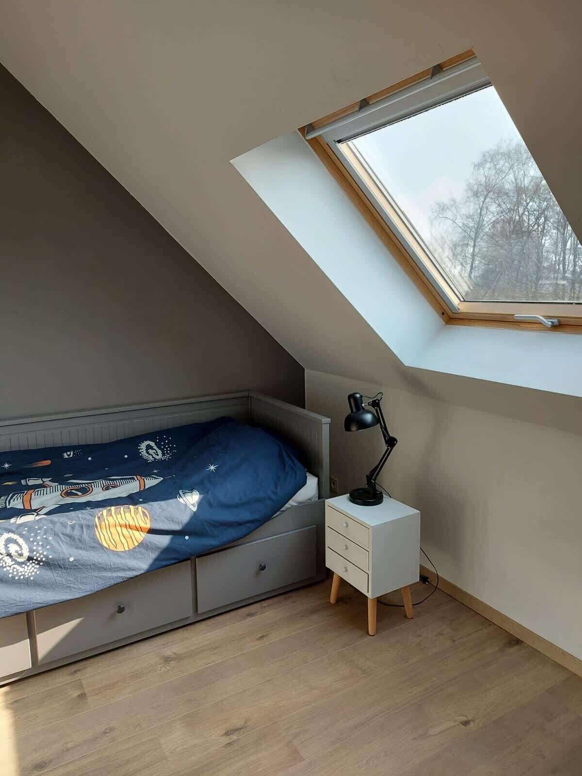 A bedroom with a sloped ceiling, a skylight, a bed with a space-themed bedspread, a bedside table with a lamp, and a small drawer unit.
