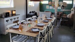 Een eetgedeelte met een lange tafel gedekt voor meerdere personen, een hangende lamp en een groene wand met diverse borden.