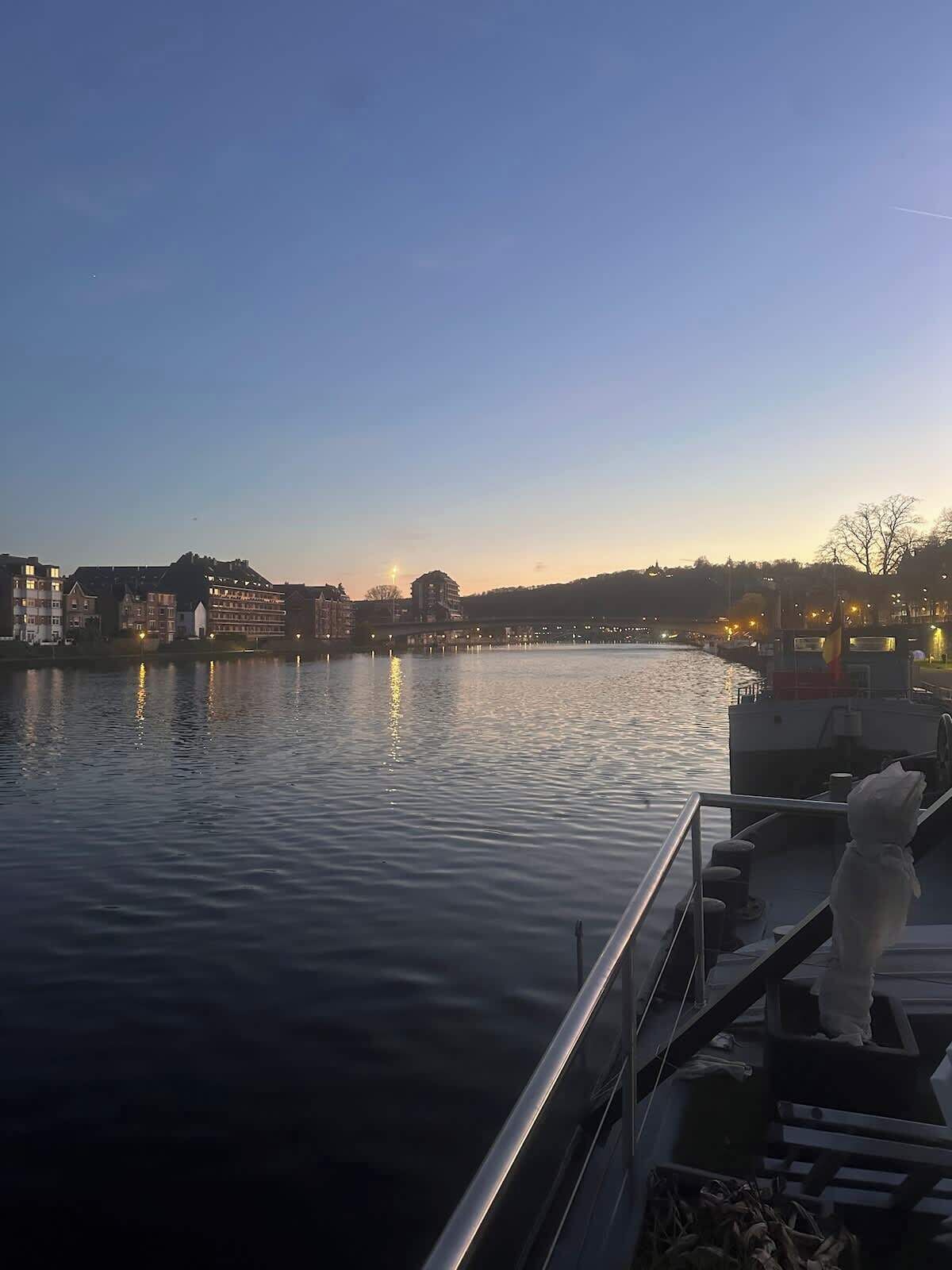 Een rustige rivier met gebouwen aan de overkant, een boot afgemeerd aan de rechterkant, en een standbeeld op het dek.