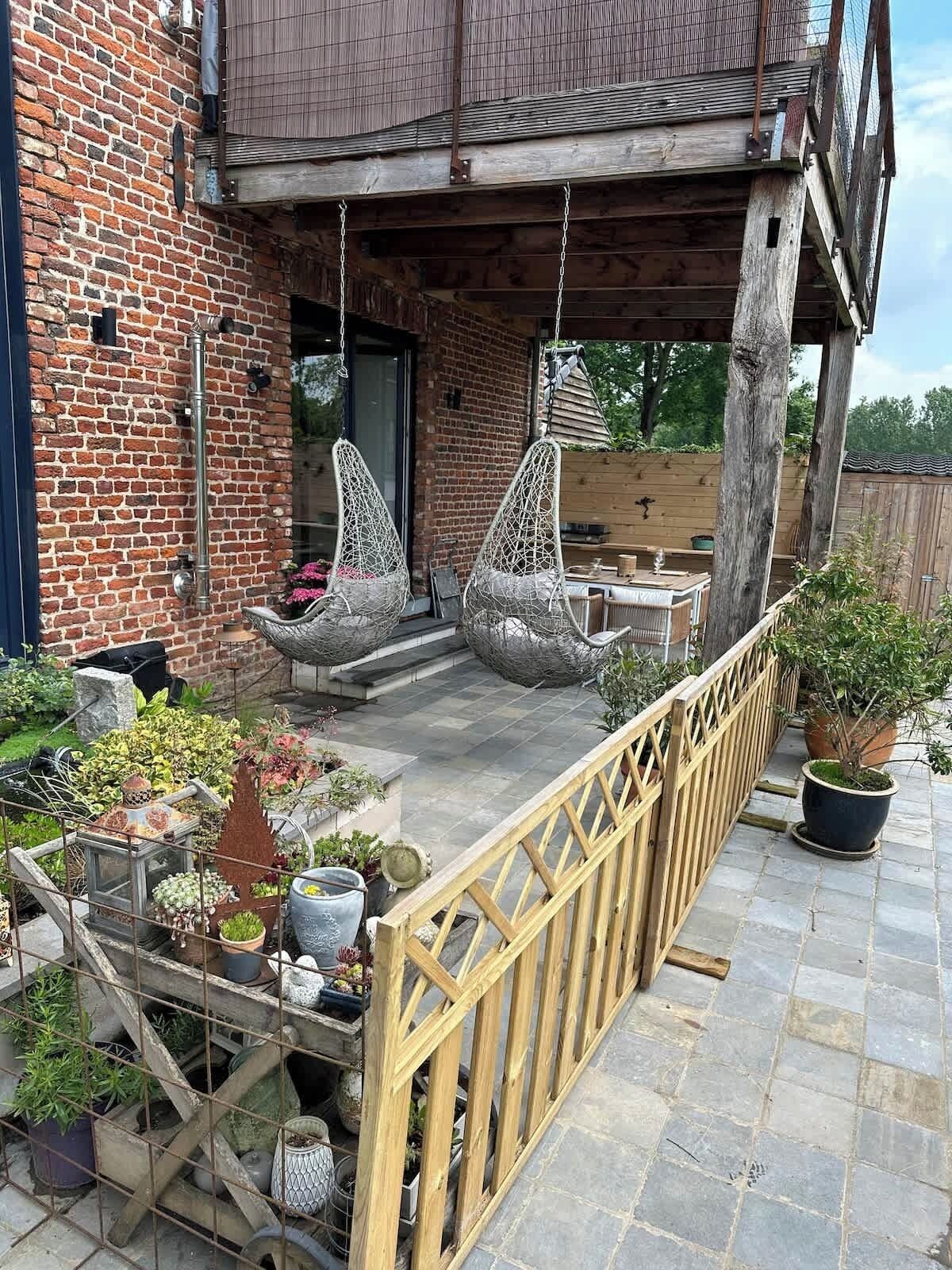 A brick building with a wooden deck, two hanging chairs, and various potted plants.