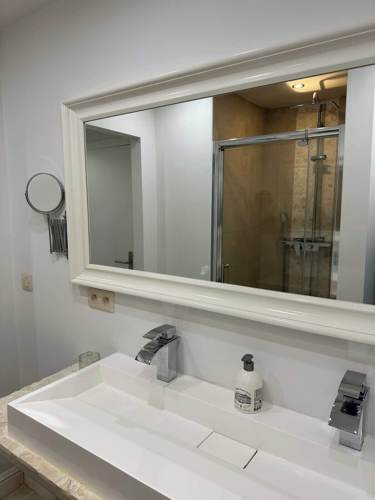 A bathroom with a white sink, a large mirror, a handheld showerhead, and a bottle of shampoo.