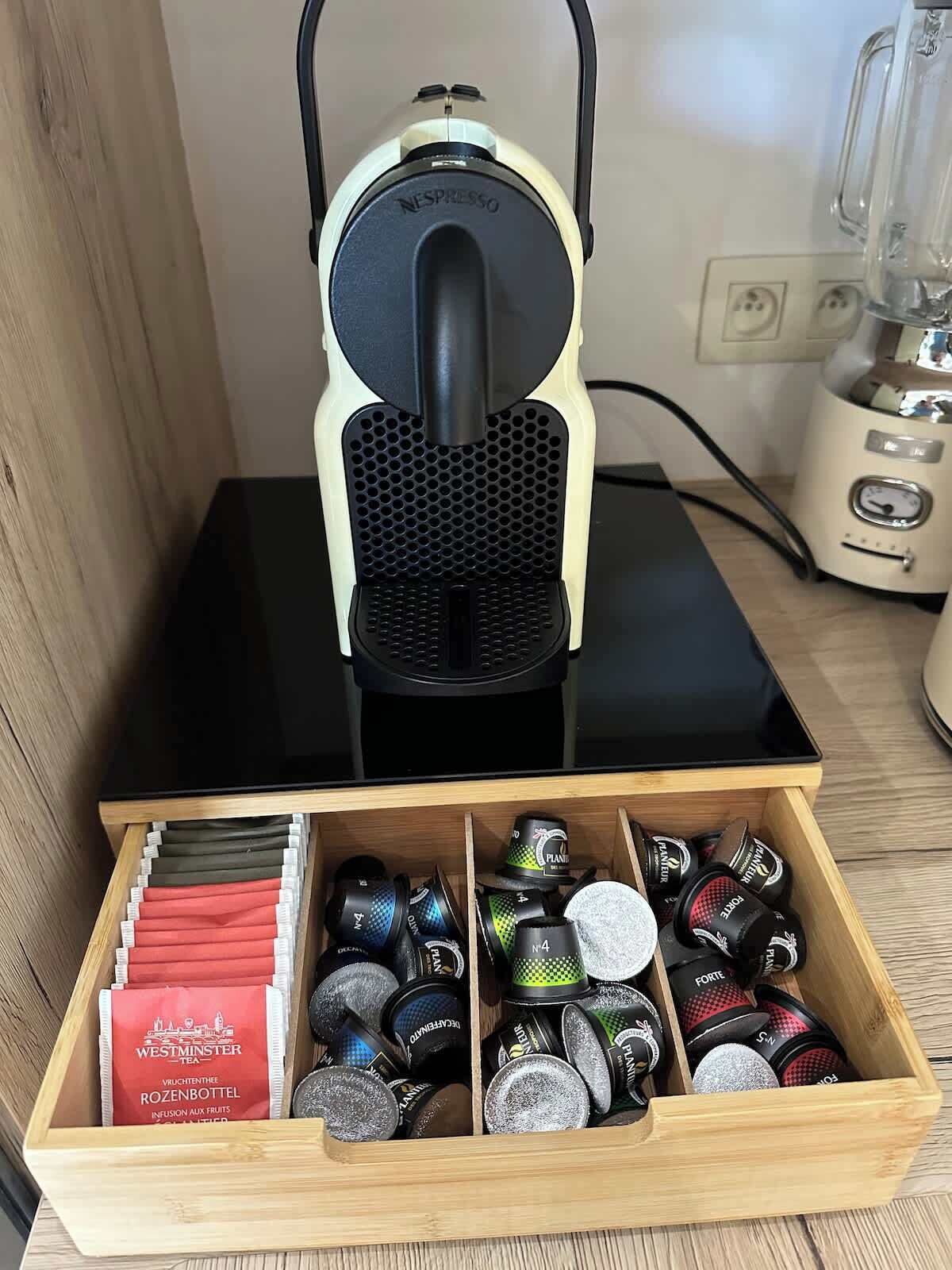 Una macchina da caffè Nespresso su un piano di lavoro nero con un cassetto sottostante contenente diverse capsule di caffè.