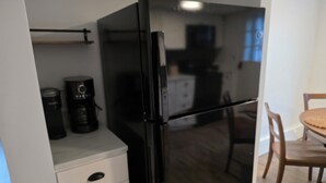 Keittiössä on musta jääkaappi, kahvinkeitin sekä ruokailutila, jossa on tuoli ja pöytä.