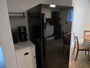 A kitchen with a black refrigerator, a coffee maker, and a dining area with a chair and table. - Hubbard Brook Getaway - King Bed - Mountain Views (Thornton)
