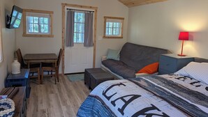 A cozy room with a wooden ceiling, a sofa, a bed with a patterned blanket, a small table, and a TV mounted on the wall. - Cozy lakefront cabin, TONS of amenities; dog friendly; 15 mins to downtown TC (Traverse City)