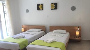 Deux lits simples, avec du linge de lit blanc et des couvre-lits verts, chacun agrémenté d’une serviette pliée posée sur le pied de lit. Des appliques murales et deux tableaux représentant des fleurs ornent le mur au-dessus des lits.