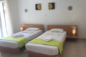 Zwei Einzelbetten mit weißen Laken und grünen Tagesdecken, jeweils mit einem auf dem Fußende gefalteten Handtuch. An der Wand sind Leuchten befestigt und über den Betten hängen zwei Blumenbilder.
