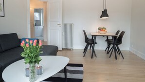 A modern living room with a sofa, a dining table with chairs, a vase with flowers, and a radiator. - Apartment Sleeps 2 With Garden (Norresundby)