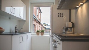 A modern kitchen with white cabinetry, a coffee machine, and a view of a brick building through a window. - Apartment Sleeps 2 With Garden (Norresundby)
