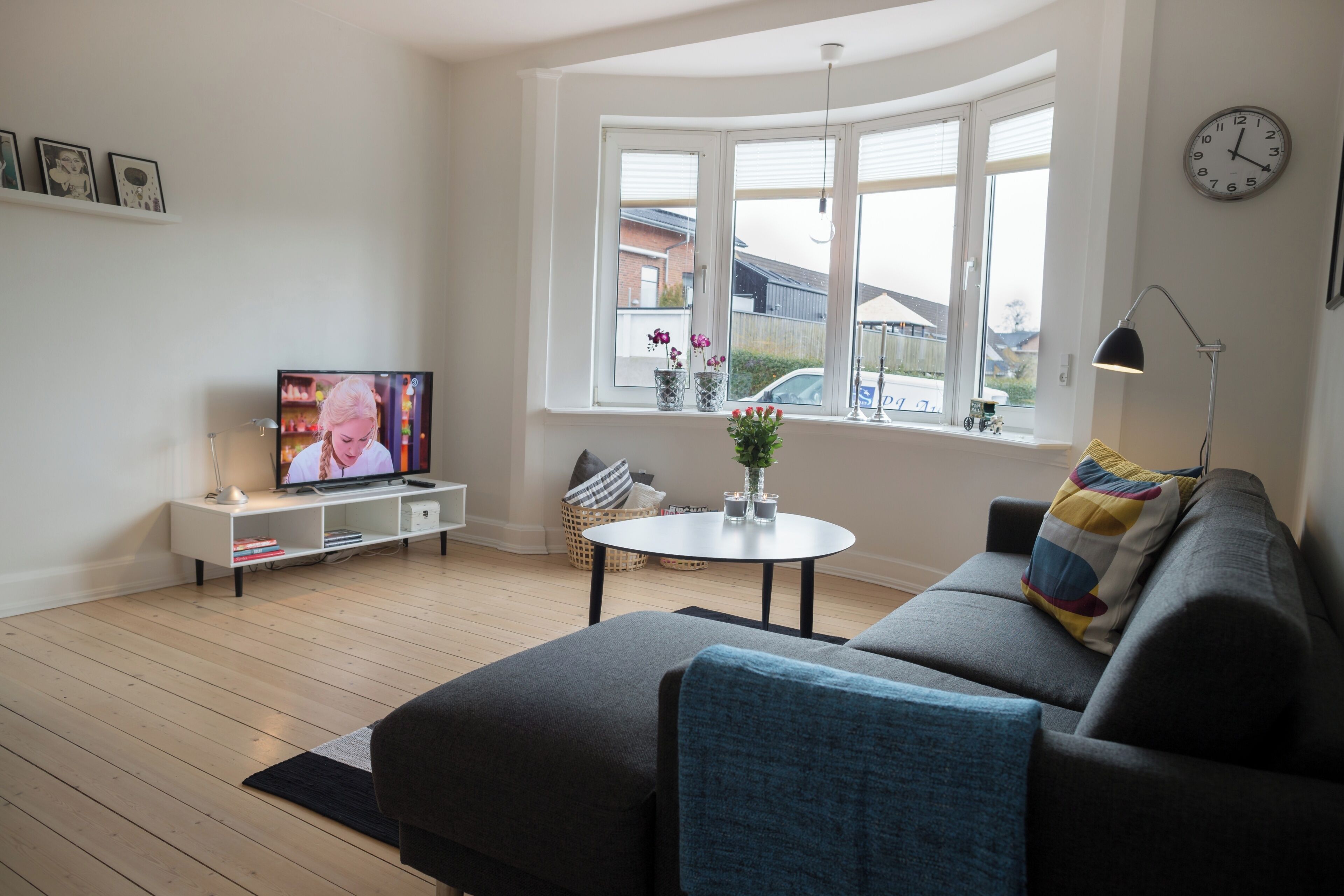 A living room with a sofa, a TV, a round table with a vase, and a window with a view of houses.