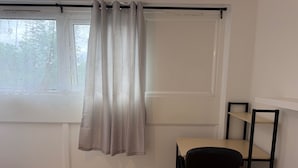 Huoneessa on ikkuna, verho, työpöytä tuolilla sekä hyllykkö.