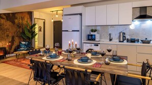 Una cocina moderna con armarios blancos, un comedor con una mesa de madera preparada para una comida, y una sala de estar con un sofá y una planta.