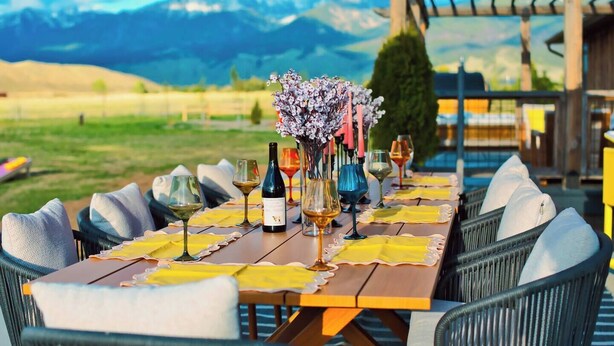 Un espace de restauration en plein air avec une table dressée pour six personnes, entourée de chaises, sous un parasol rouge, et offrant une vue sur les montagnes en arrière-plan.