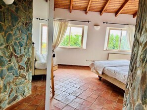 A bedroom with a stone wall, two windows with curtains, a bed, and a wooden ceiling.