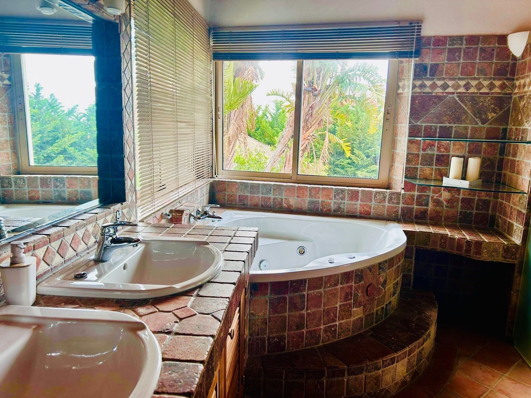 Une salle de bain avec une baignoire, deux lavabos, une fenêtre donnant sur un espace verdoyant et un mur carrelé.