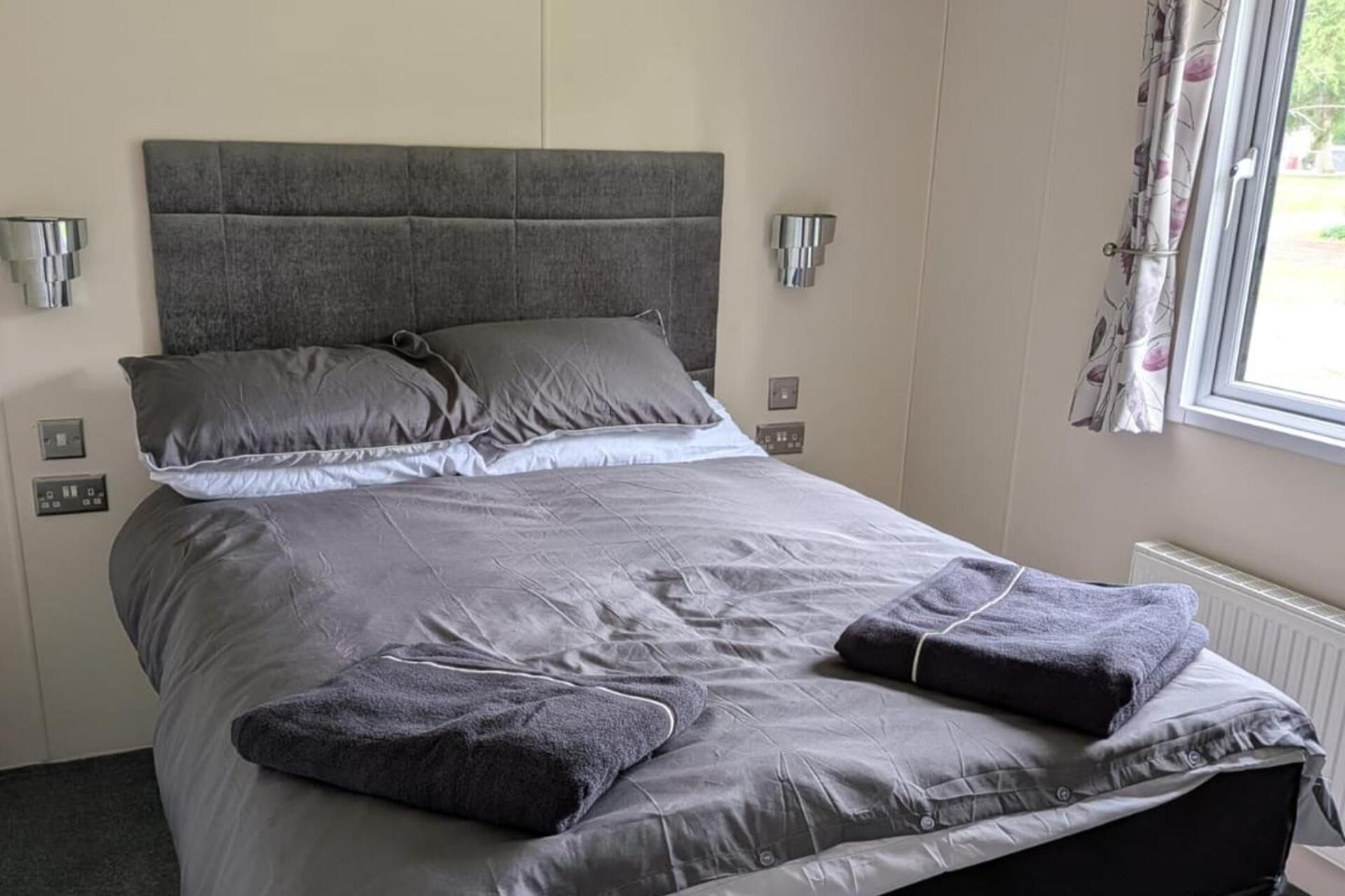 Ein ordentlich bezogenes Bett mit grauem Bettzeug und Kissen, ein grauer Kopfteil und ein Fenster mit Vorhängen.