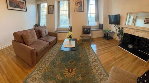 Ein Wohnzimmer mit braunem Sofa, einem kleinen Tisch mit Blumenvase, einem gemusterten Teppich, einem Kamin und einem Fernseher.