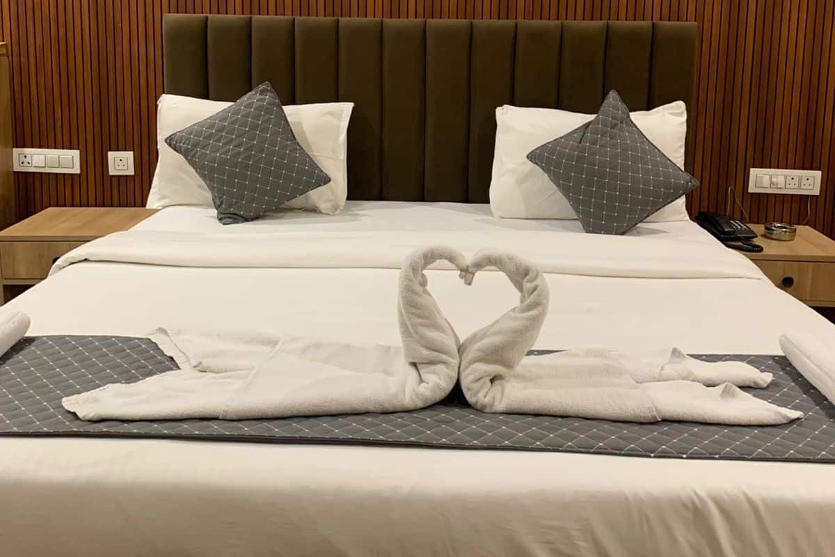 Habitación de hotel con pared de paneles de madera, cama con ropa de cama blanca, dos almohadas, una disposición de toallas con forma de corazón y una mesita de noche con teléfono y lámpara.