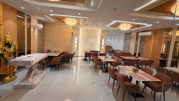 A hotel dining area with multiple tables and chairs, a large chandelier, and a statue on the left. - Hotel Tulsi Tower (Raipur)