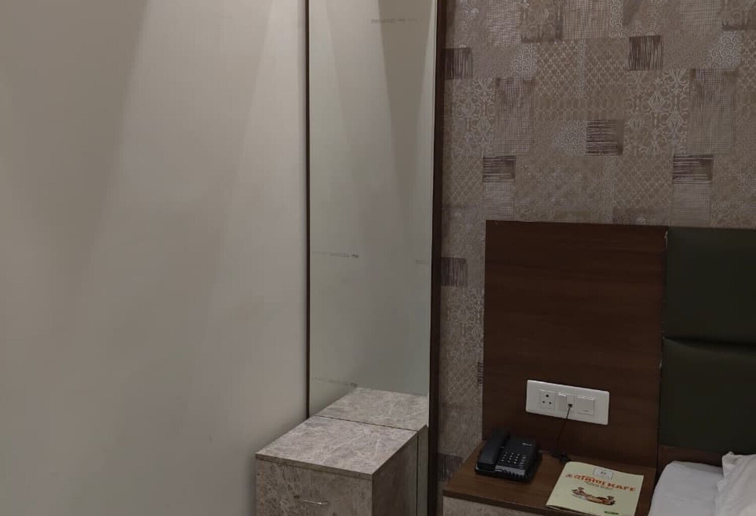 Habitación de hotel con cama, mesita de noche con teléfono y menú, interruptor de luz en la pared y un armario pequeño.