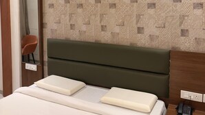 A hotel room with a patterned wallpaper, a green upholstered headboard, a white bed with two pillows, a wooden nightstand with a phone, and a mirror on the wall. - Hotel Tulsi Tower (Raipur)
