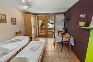 A room with two single beds, a small table with chairs, a window with a view of greenery, and a door leading outside. - Convenient studio with view in the center of the resort (Anniviers)