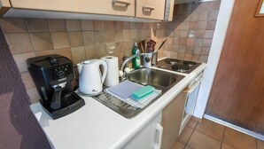 Et kompakt køkken med kaffemaskine, elkedel, vask og diverse køkkenredskaber.