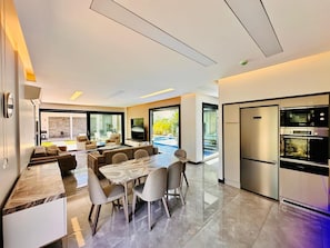 Una cocina moderna con horno empotrado y microondas, un área de comedor con mesa y sillas de mármol, y un amplio ventanal con vista a una piscina.