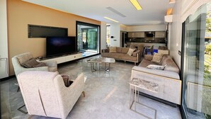 A modern living room with a flat-screen TV, comfortable seating, a glass coffee table, and a view of the outdoors.