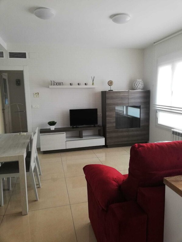 Un soggiorno moderno con TV, un divano rosso, un'area pranzo con tavolo e sedie, e un angolo cottura con mobili e una finestra.