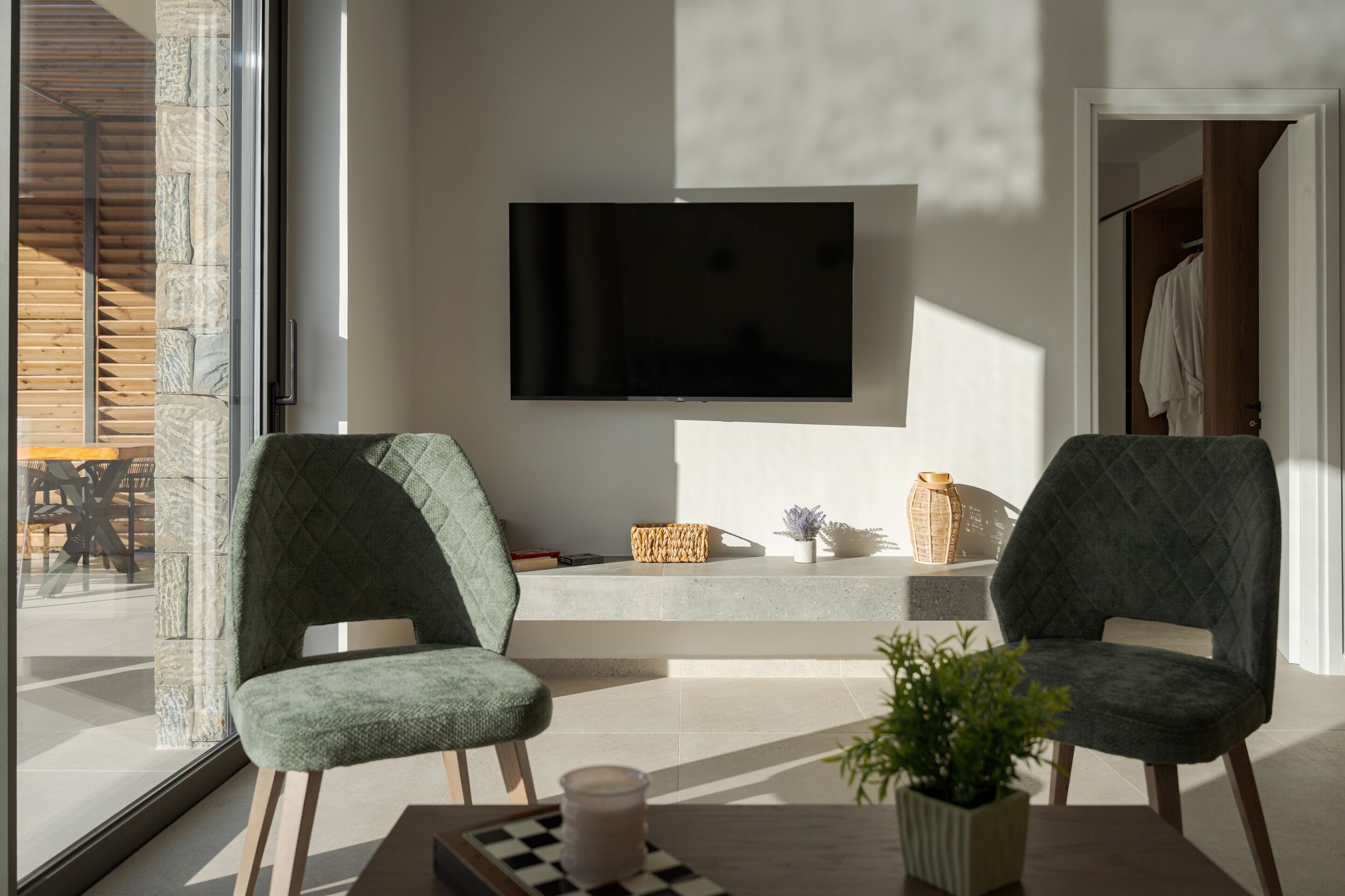 Egy modern nappali két zöld színű, kárpitozott székkel, egy síkképernyős televízióval, egy kis asztallal, melyen egy gyertya és egy sakkjátszma található, valamint egy virágos növényt tartalmazó cseréppel.