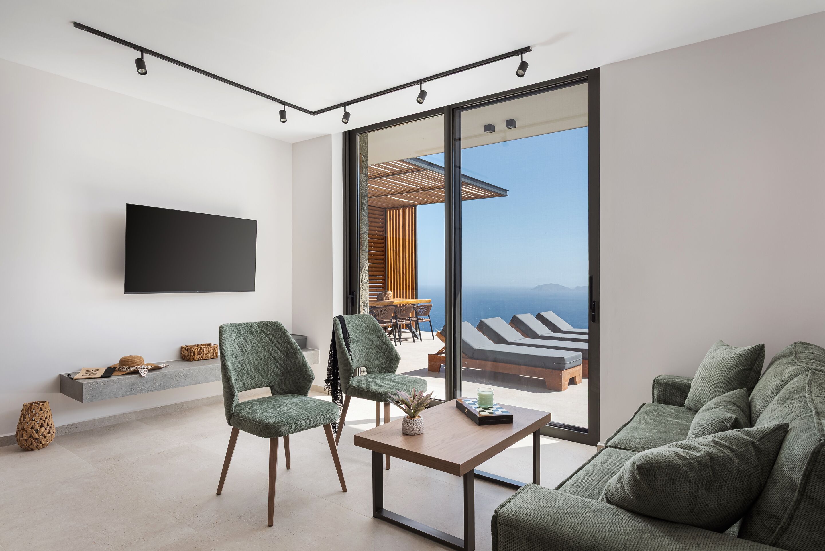 A modern living room with a flat-screen TV, green upholstered furniture, a wooden coffee table, and a large sliding glass door leading to a balcony with a view of the ocean.