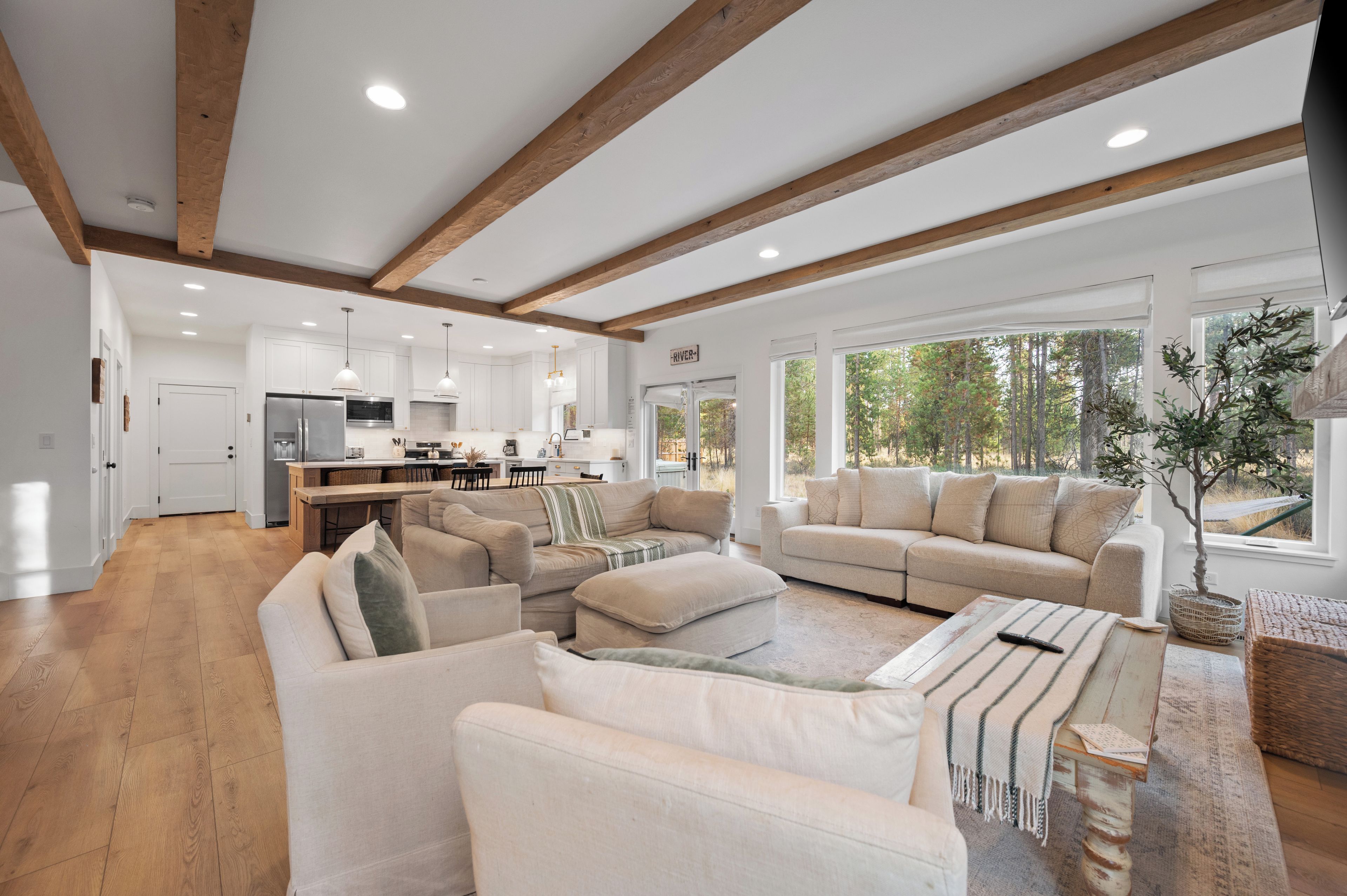 A spacious living area with a large sofa, a coffee table, and a dining table. There is a kitchen area with white cabinets and a dining table. The room has wooden beams on the ceiling and large windows with a view of trees.