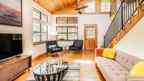 A spacious living room with a large TV, two leather armchairs, a glass coffee table, and a staircase with a wooden railing. - Olinca Ranch - Private Texas Oasis (Driftwood)