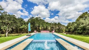 A rectangular pool with a water fountain in the center, surrounded by a paved deck. There are lounge chairs and umbrellas along the poolside. - Olinca Ranch - Private Texas Oasis (Driftwood)
