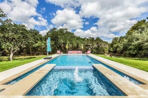 Ein rechteckiger Pool mit einer Fontäne in der Mitte, umgeben von einer gepflasterten Liegefläche. Am Poolufer befinden sich Liegestühle und Sonnenschirme.