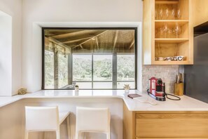 A modern kitchen with a large window, a coffee machine, and a wooden shelf with glasses. - Olinca Ranch (Driftwood)