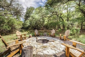 A stone fire pit surrounded by wooden Adirondack chairs in a forested area. - Olinca Ranch (Driftwood)