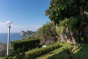 Eine Küstenlandschaft mit freiem Blick auf das Meer, einer weißen, kugelförmigen Leuchte und verschiedenen Grünpflanzen, darunter eine Palme und ein Heckenstreifen.