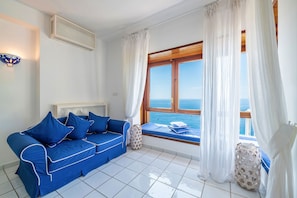 Ein Zimmer mit einem blauen Sofa, einem Fenster mit Meerblick und einem weiß gefliesten Boden.