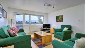 Una sala de estar moderna con sofás verdes, una mesa de centro de madera, un televisor de pantalla plana y un amplio ventanal con vista a la ciudad.