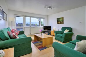 Ein modernes Wohnzimmer mit grünen Sofas, einem Holztisch, einem Flachbildfernseher und einem großen Fenster mit Blick auf die Stadt.